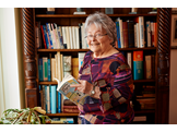 Mimi Reed smiles and holds a book, standing in front of a bookshelf in her home.